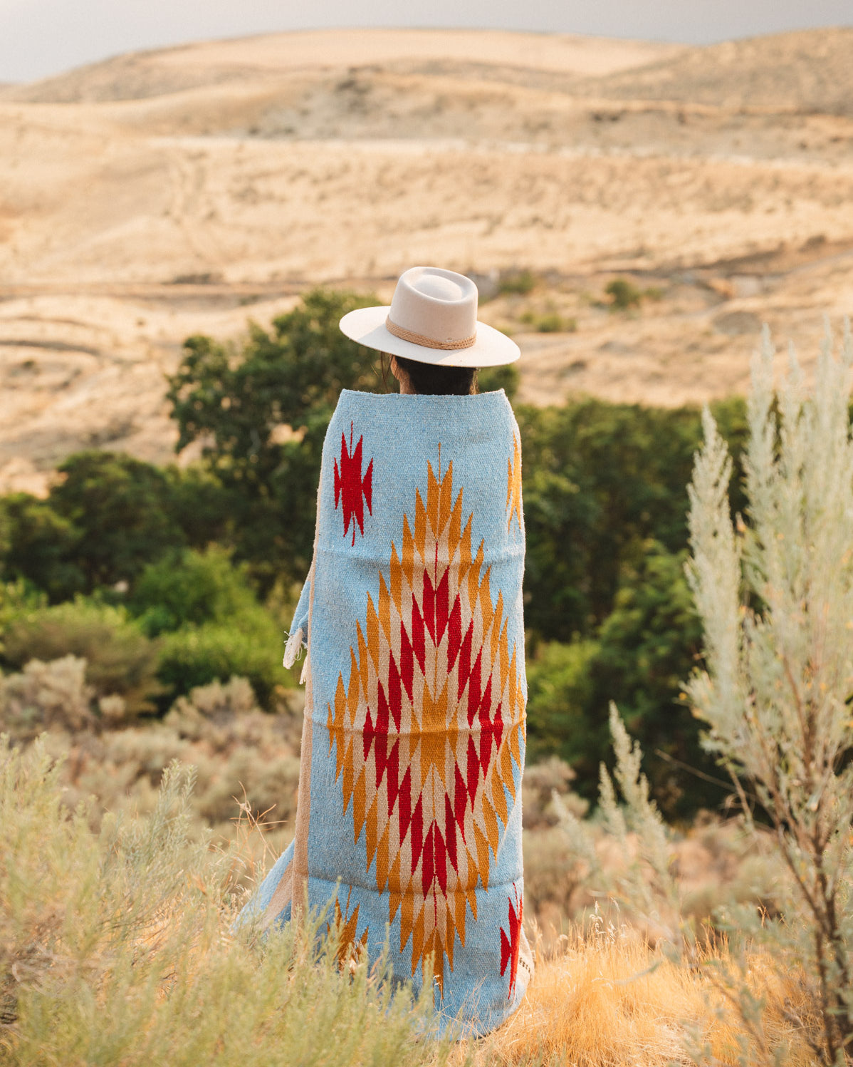 A person standing outdoors in a desert landscape, viewed from behind, wrapped in a light blue blanket with a large red, orange, and yellow geometric diamond pattern and wearing a white hat.