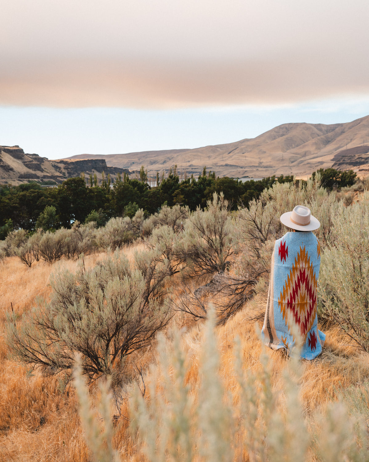 A person stands in a dry grassy landscape wearing a wide-brimmed hat and a light blue blanket or poncho with a large red, yellow, and white geometric diamond pattern, with hills and trees in the background.