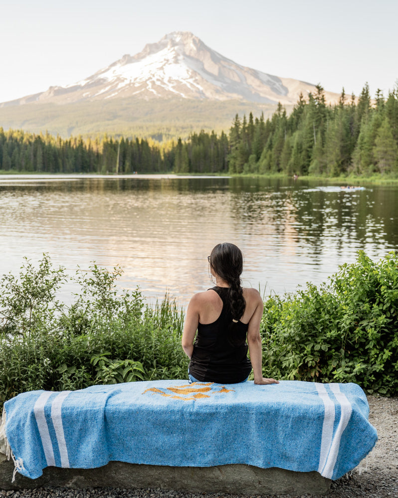 Woman sitting on blue blanket by a lake and mountain at sunset