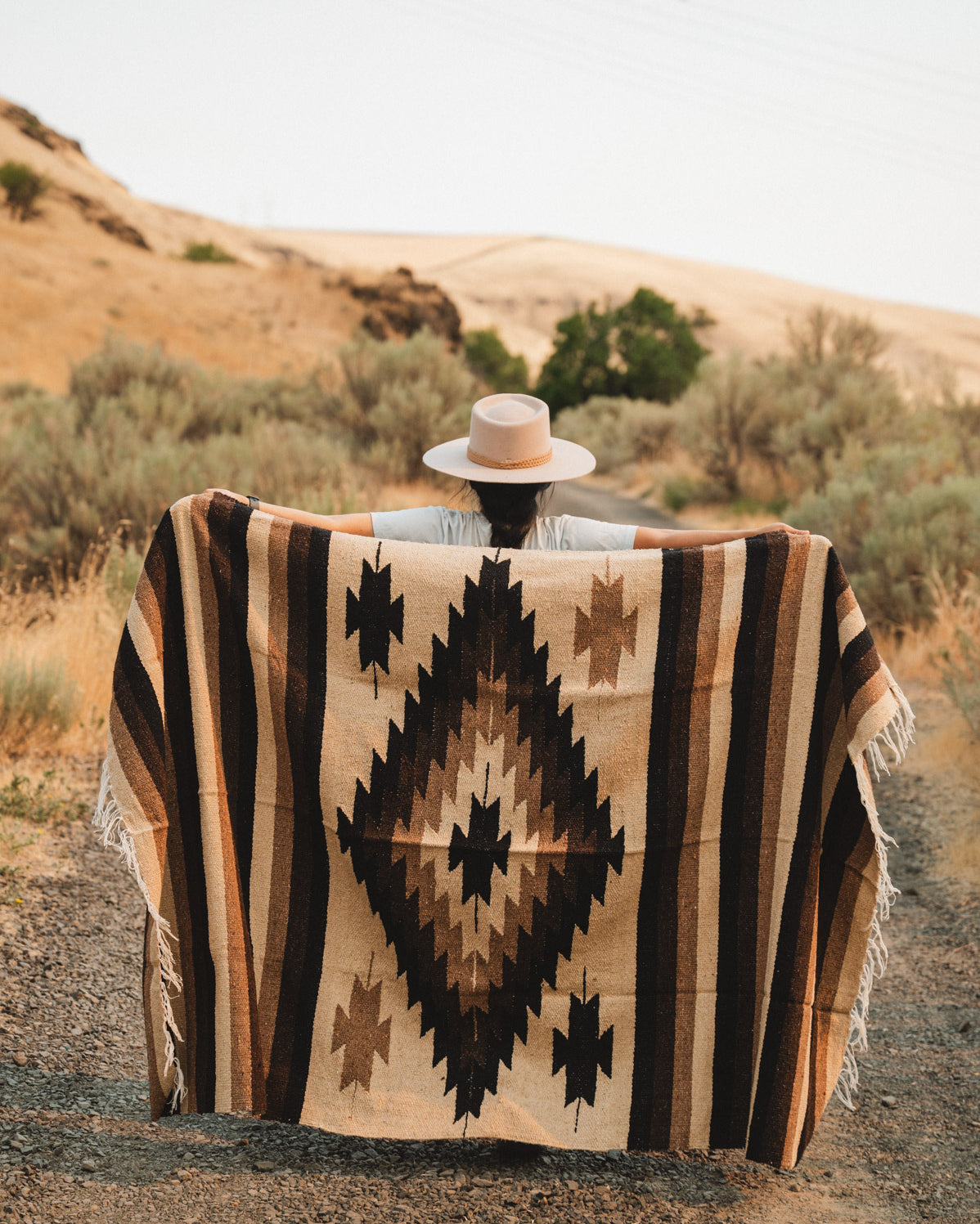 A person standing outdoors on a dirt path in a desert landscape, holding up a large woven blanket or poncho with a Southwestern geometric pattern in beige, brown, and black. The person is wearing a wide-brimmed hat.