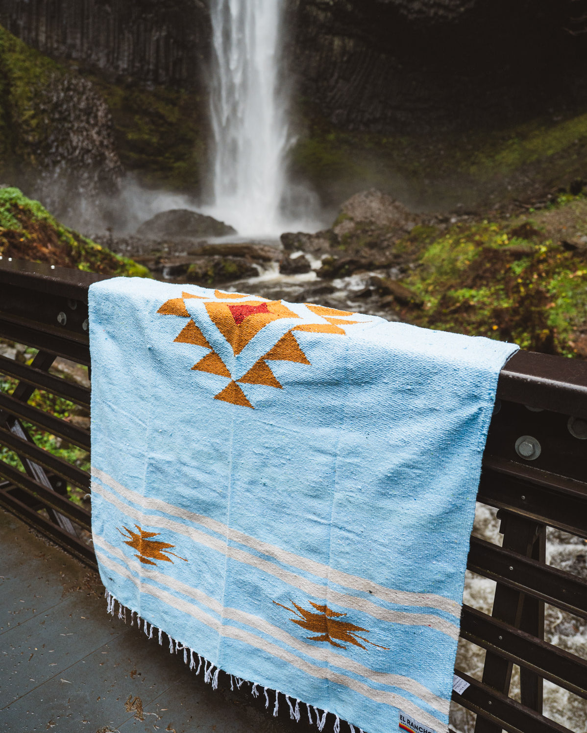 Blue quilt with orange and yellow patterns draped over a railing in front of a waterfall.