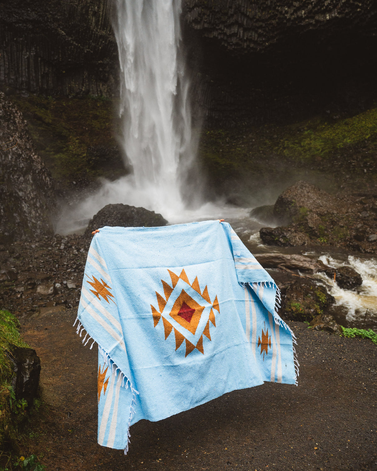 Light blue blanket with a geometric pattern draped over a rock in front of a waterfall.