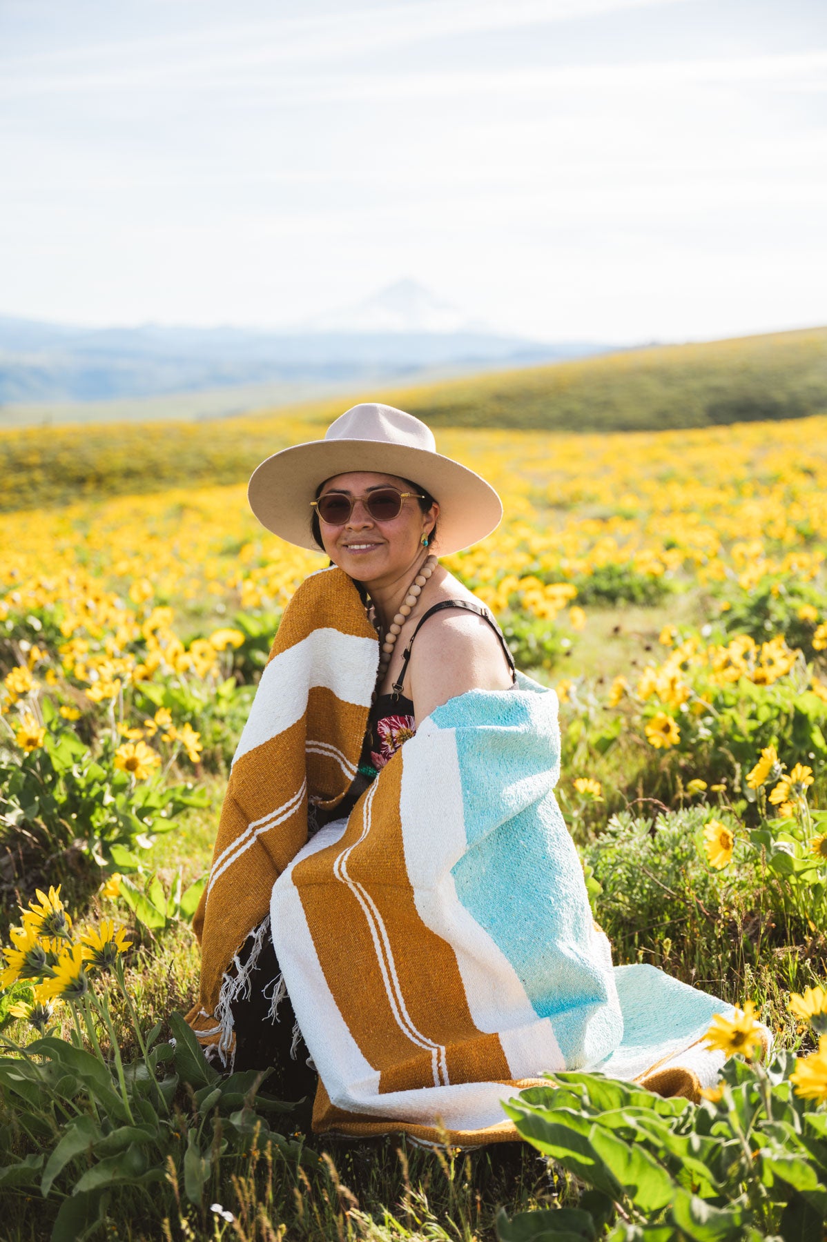 Person sitting in a field of yellow flowers wearing a wide-brimmed hat and sunglasses, with a colorful towel draped over their shoulders.