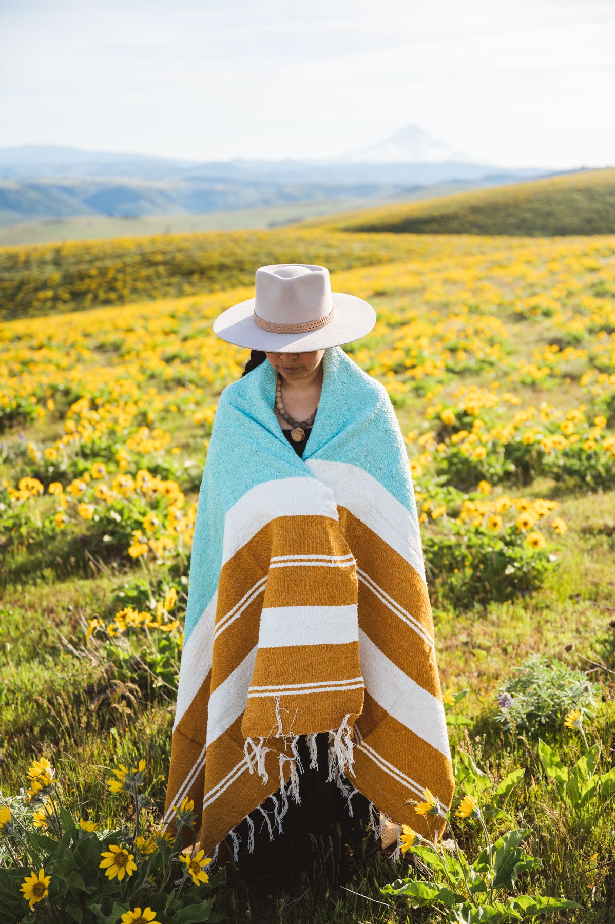 Person wrapped in a striped towel standing in a field of yellow flowers with mountains in the background