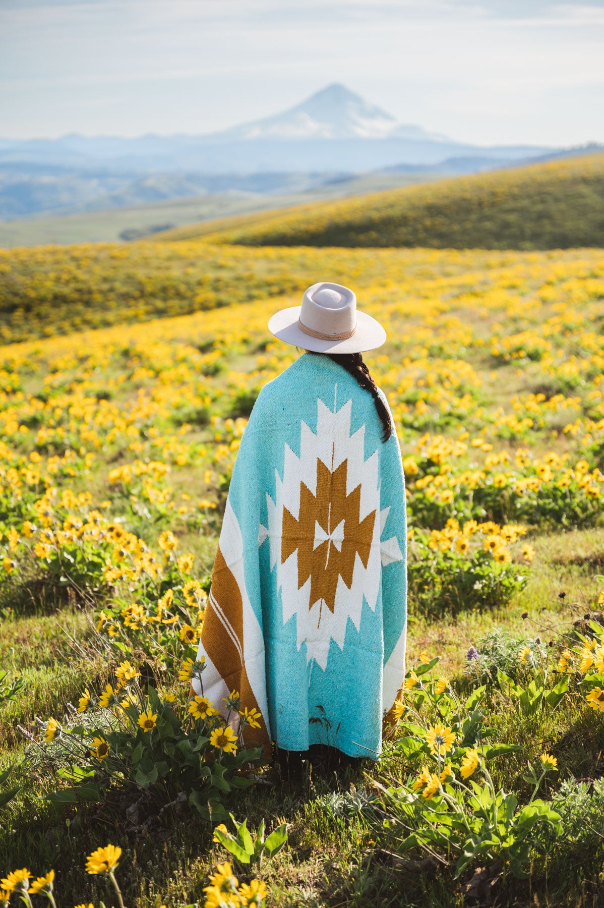 Person wearing a turquoise blanket with a white and brown diamond pattern in a field of yellow flowers with mountains in the background.