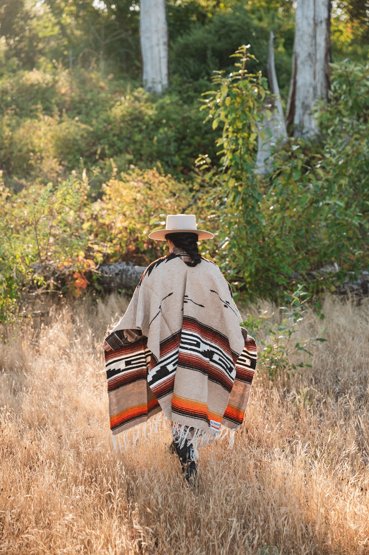 A person viewed from behind walking through tall dry grass in a wooded outdoor setting, wearing a wide-brim hat and a beige woven poncho with black, white, and orange geometric pattern details.