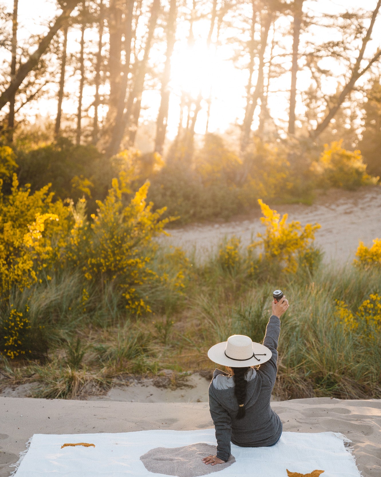 Woman holding a beer sitting on cream blanket in the sand at sunset