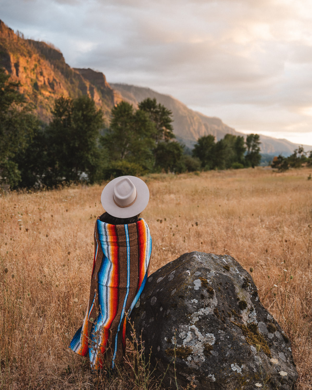 Woman standing next to rock in field wrapped in handwoven brown saltillo blanket
