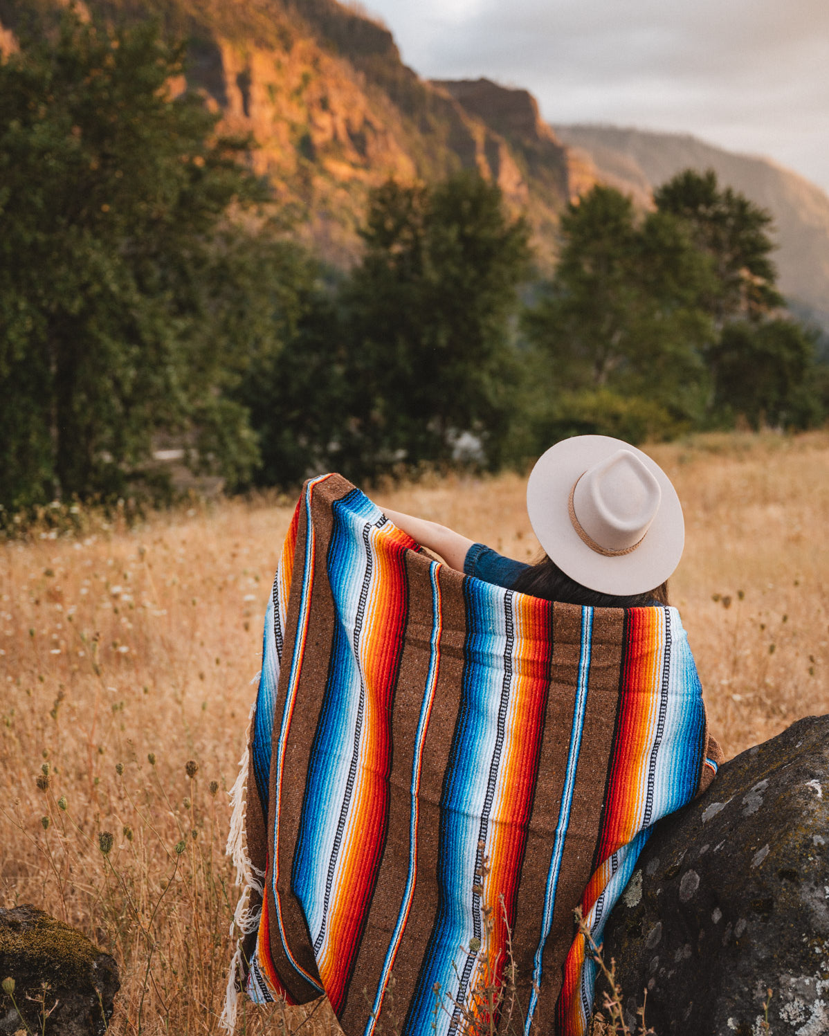 Woman standing next to rock in field wrapped in handwoven brown saltillo blanket