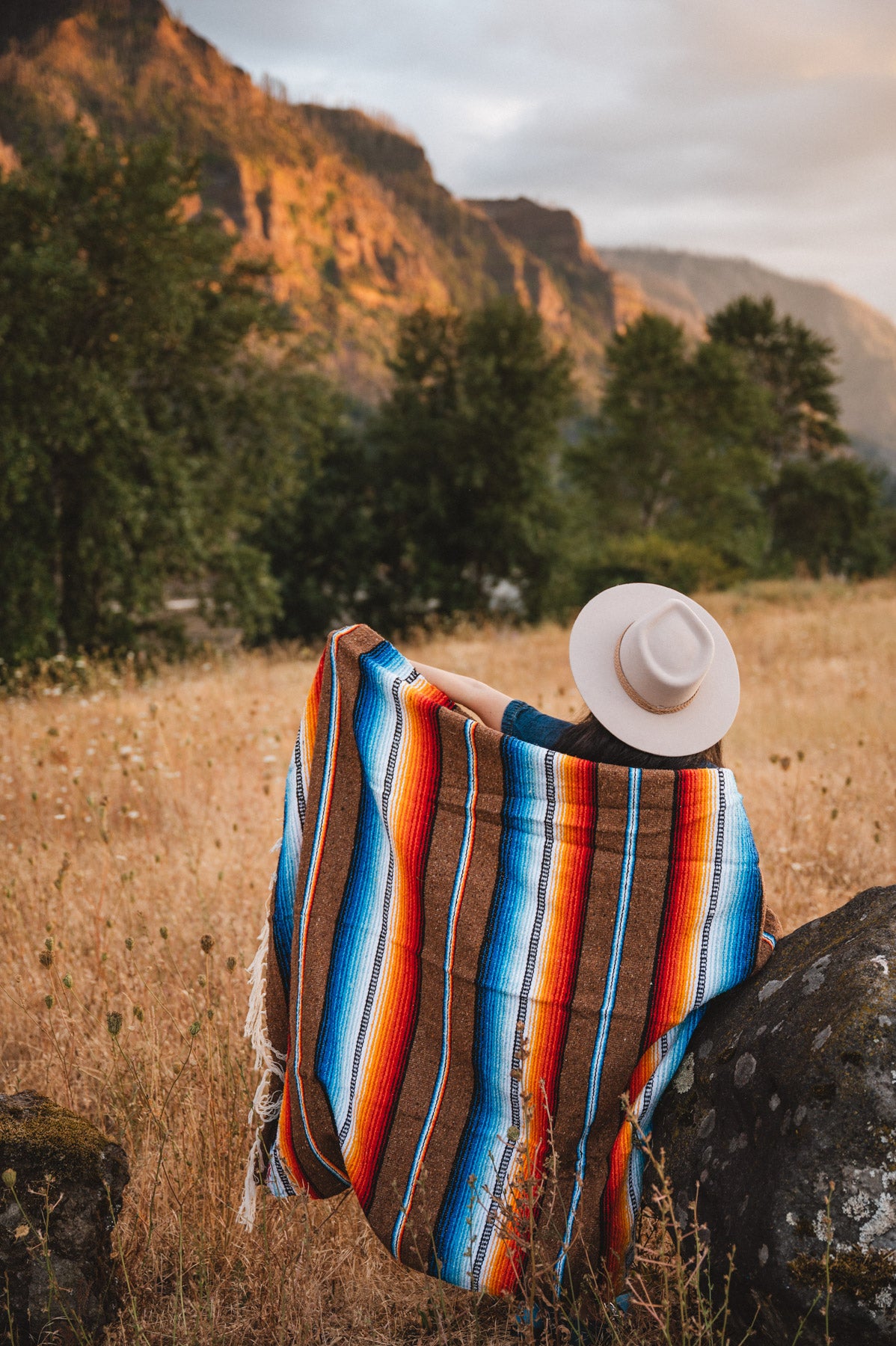 Woman standing next to rock in field wrapped in handwoven brown saltillo blanket