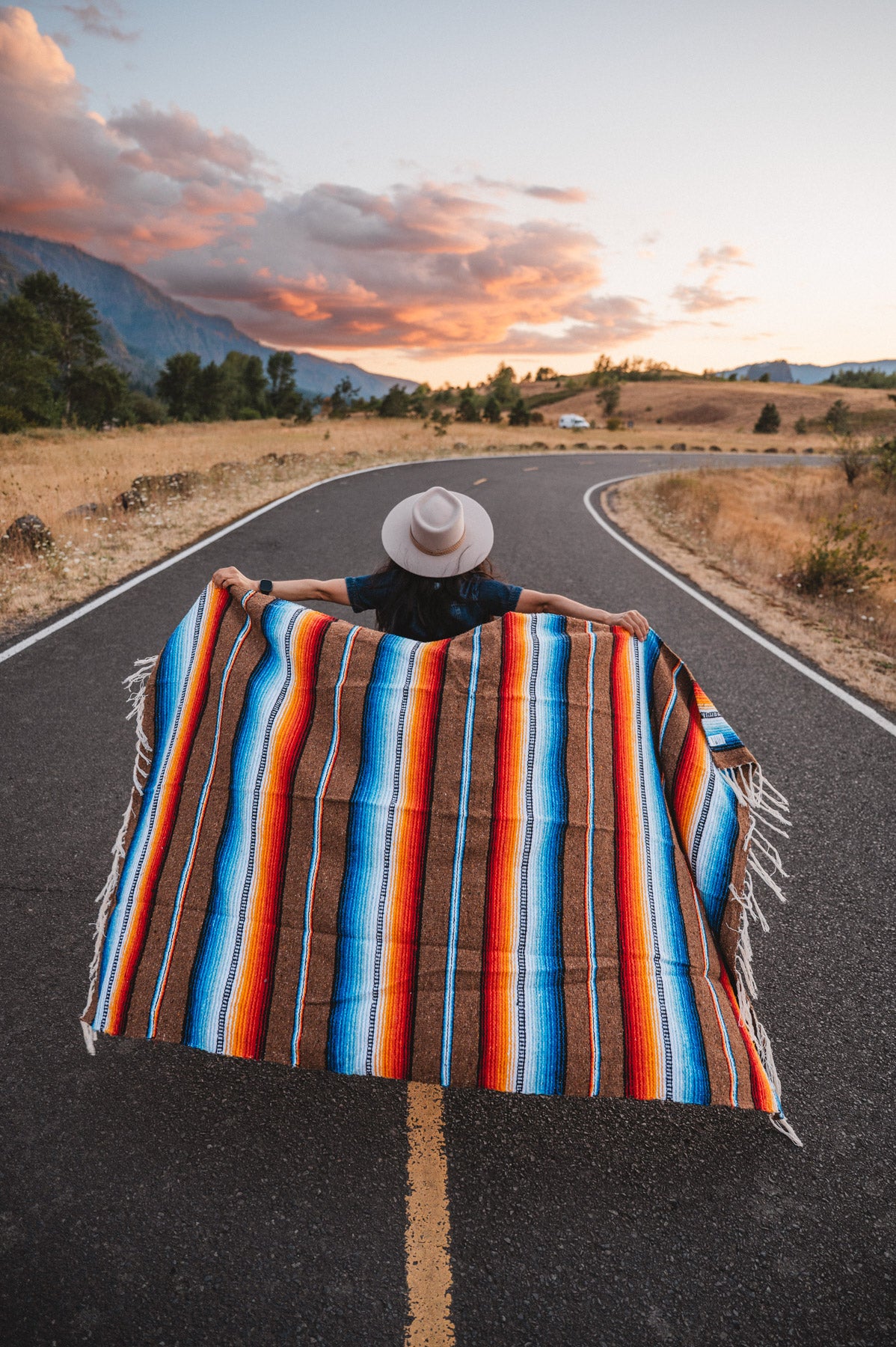 Woman walking down road holding handowoven saltillo blanket behind her at sunset