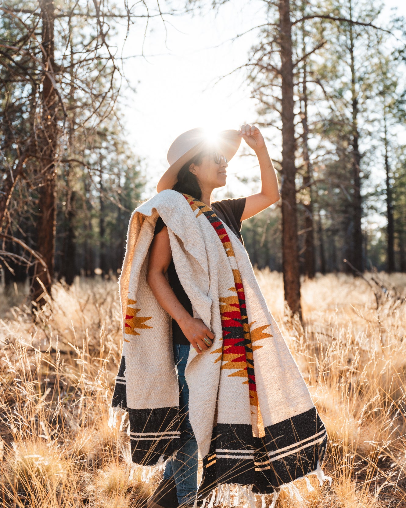 Woman wearing cowboy hat standing in a dry grassy field and forest at sunset wearing a southwestern diamond blanket over her shoulder