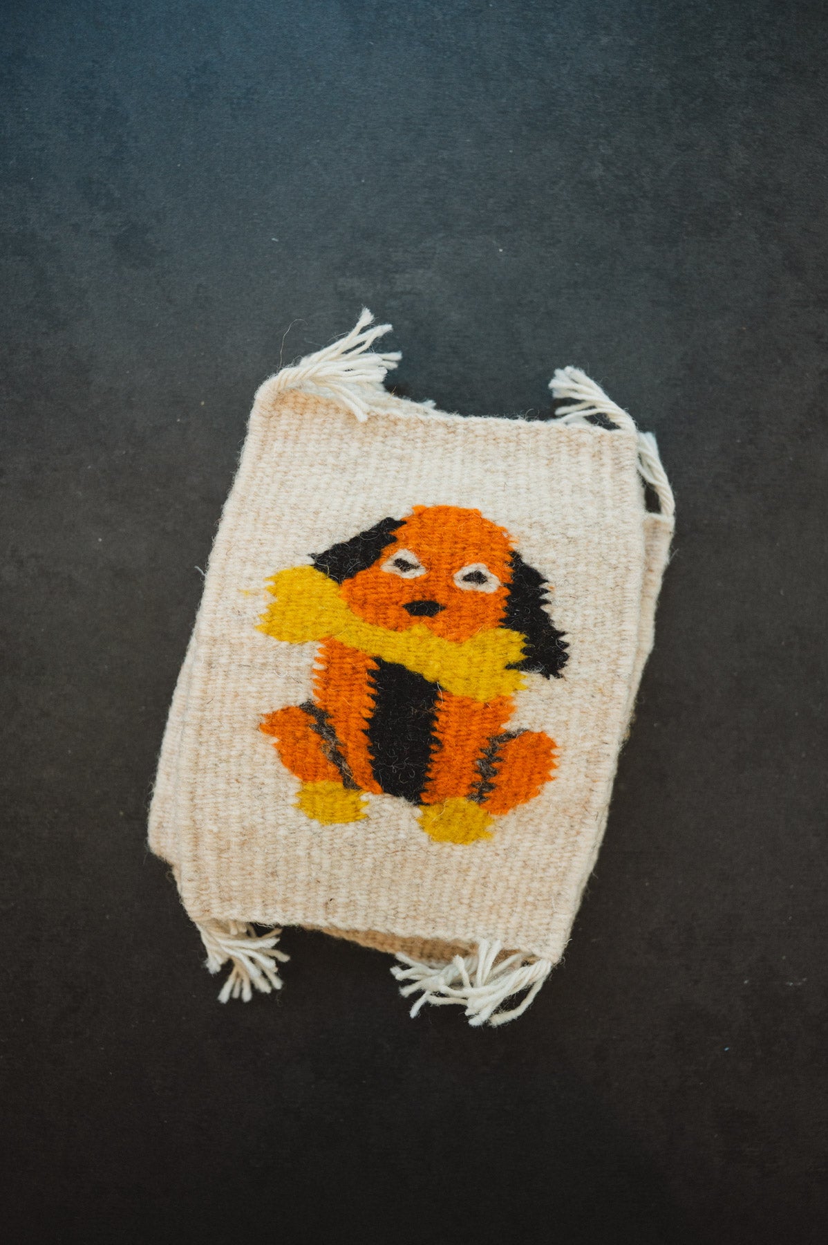 A small woven or knitted beige pouch with frayed edges, featuring a colorful dog motif in orange, black, and yellow on the front, photographed against a dark textured background.