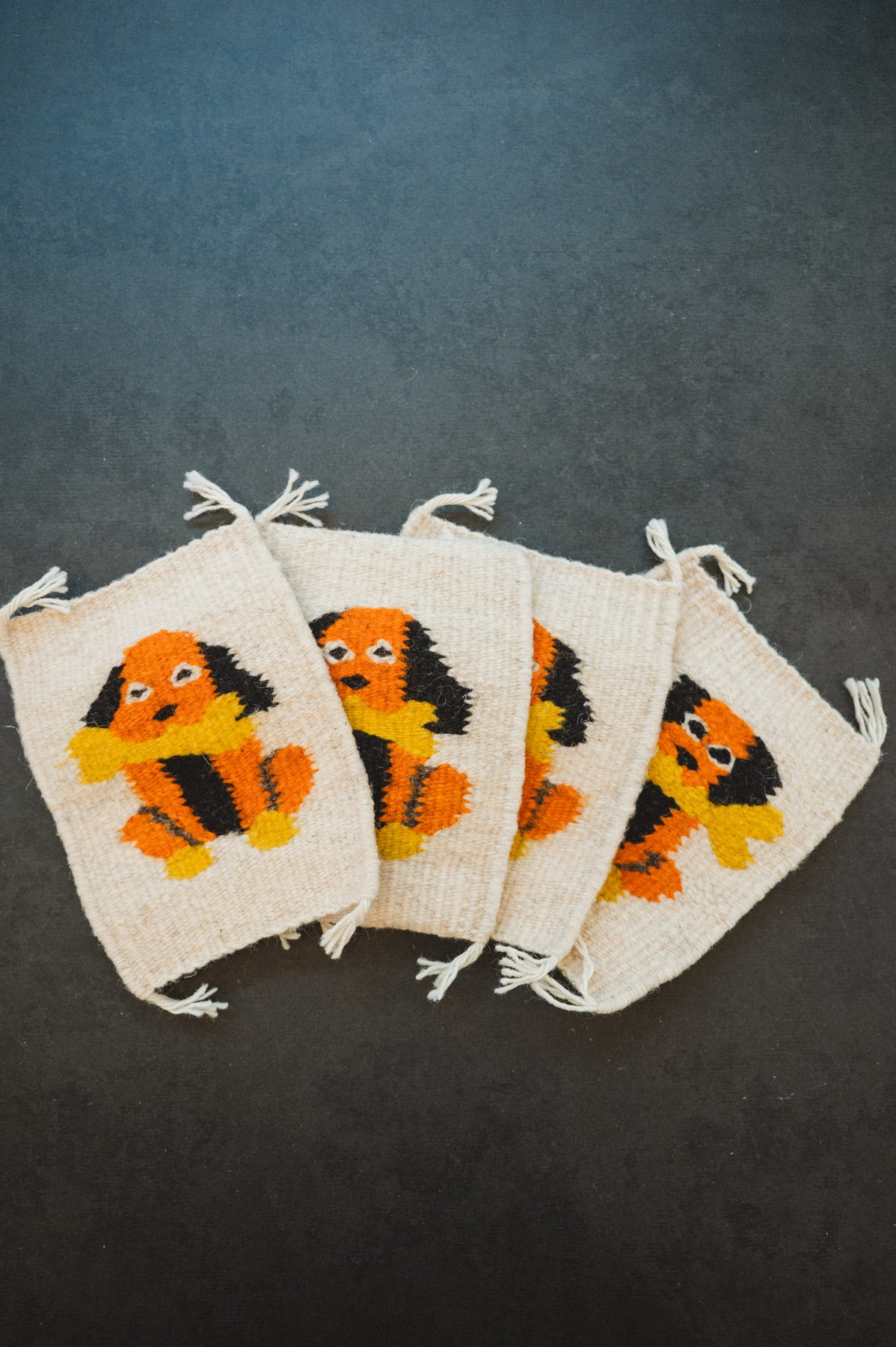 Four cream-colored woven drawstring pouches with a colorful embroidered dog motif in orange, black, and yellow, arranged on a dark gray background.