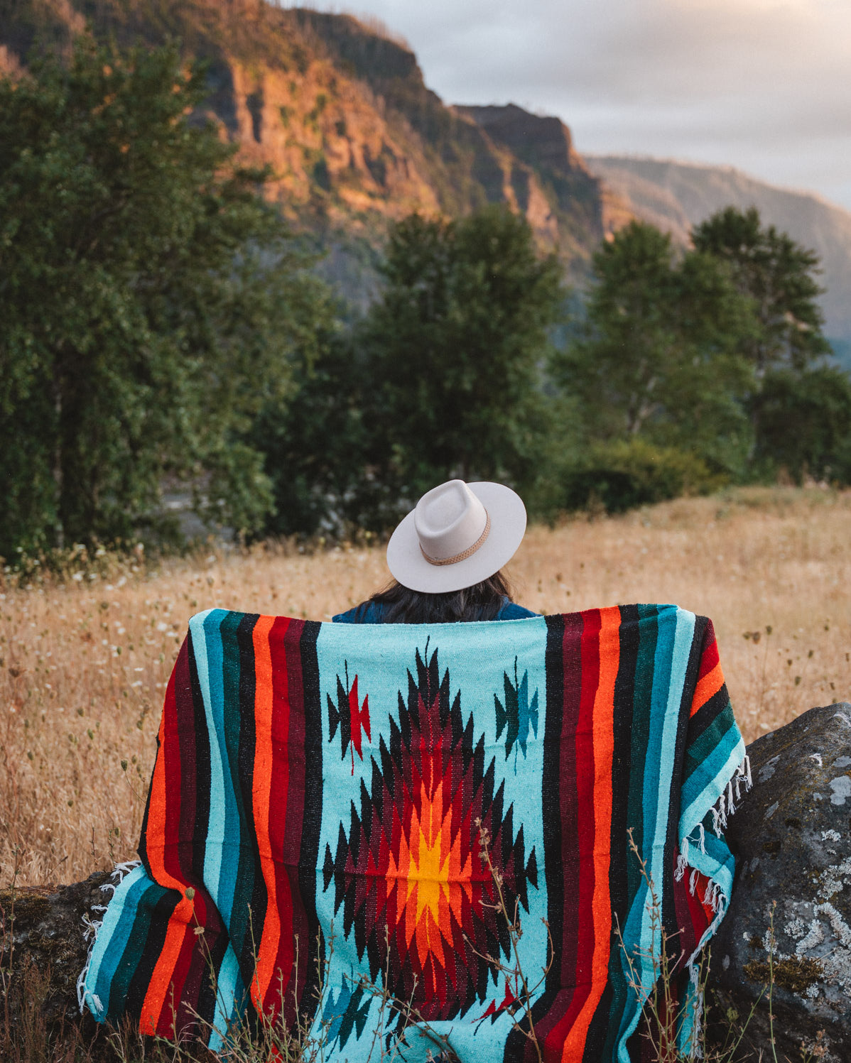 A person sitting outdoors in a grassy field with mountains and trees in the background, viewed from behind while wearing a white wide-brim hat and wrapped in a colorful Southwestern-style woven blanket with turquoise, red, orange, black, and cream geometric patterns.
