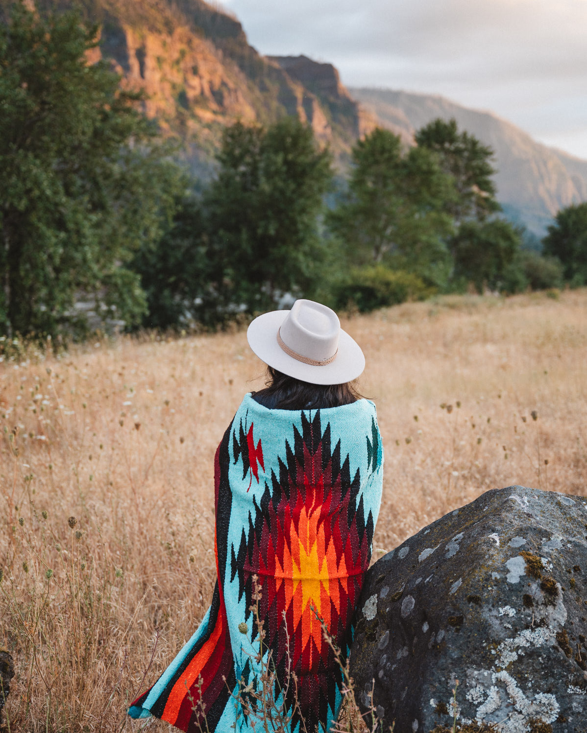 A person seen from behind sitting in a grassy field with mountains and trees in the background, wearing a white hat and a colorful southwestern-style blanket or poncho with a turquoise, red, orange, and black geometric pattern.