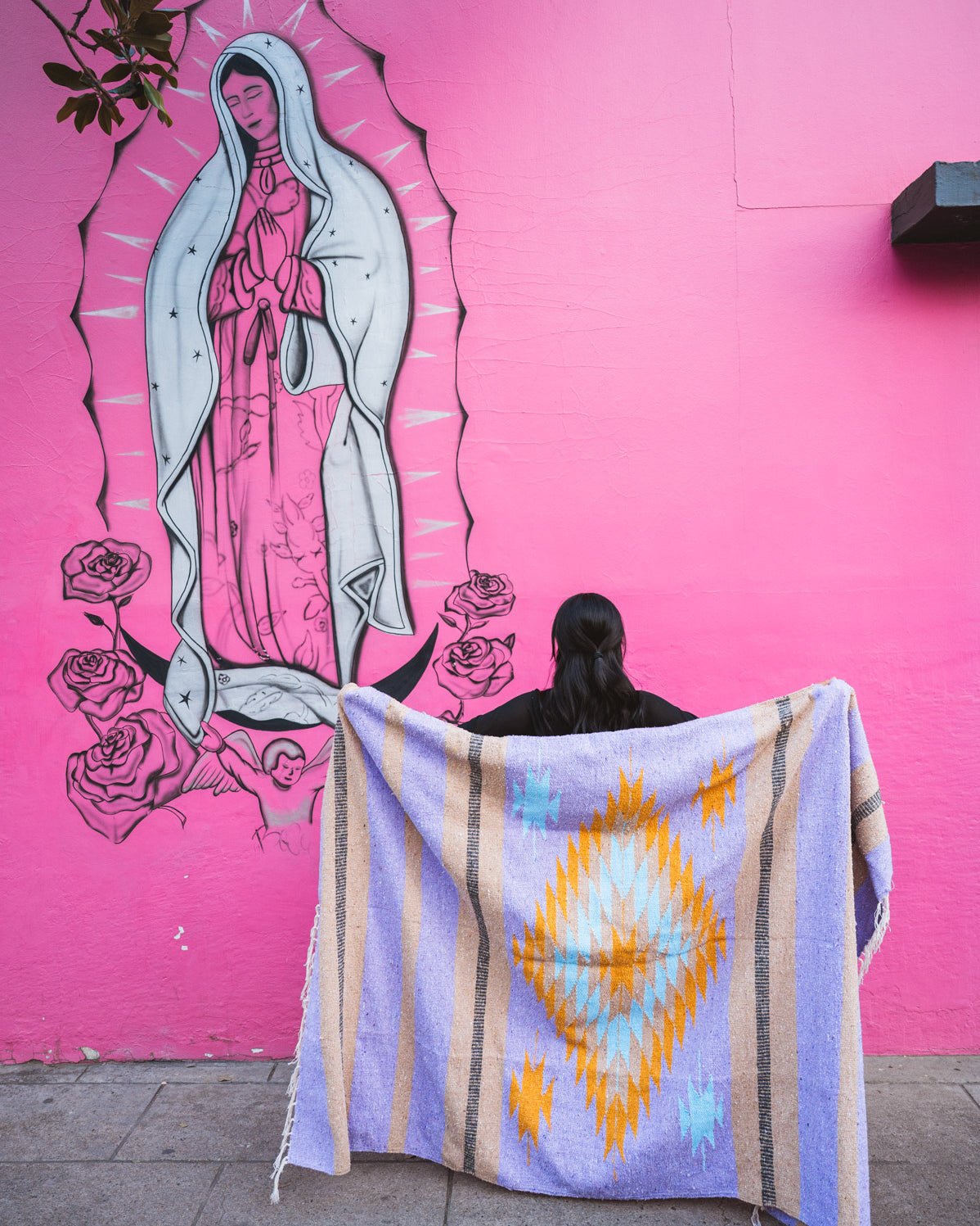 Person holding a colorful blanket in front of a pink wall with a mural of Our Lady of Guadalupe.