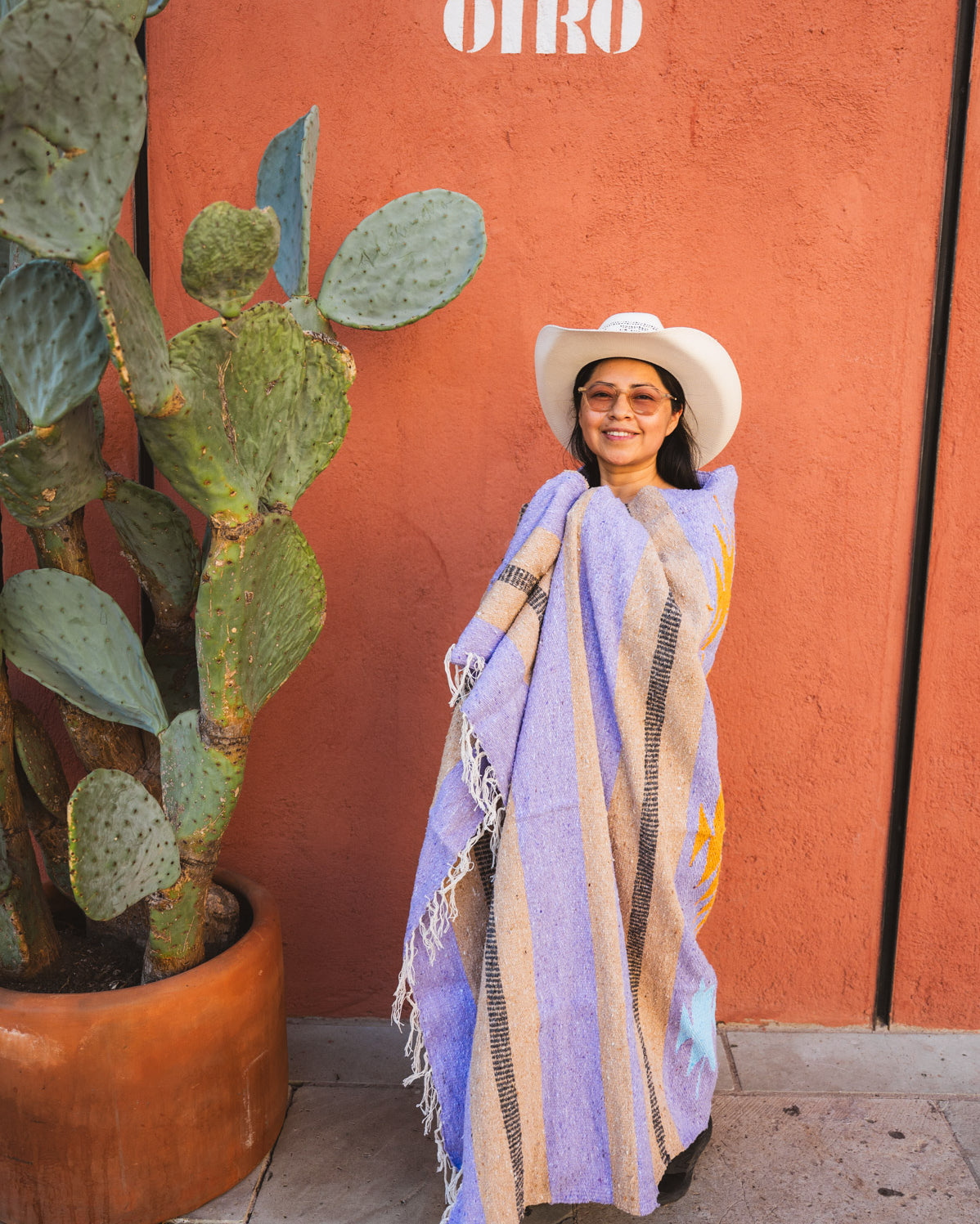 Woman in a purple shawl and white hat standing next to a cactus plant with a red wall in the background.