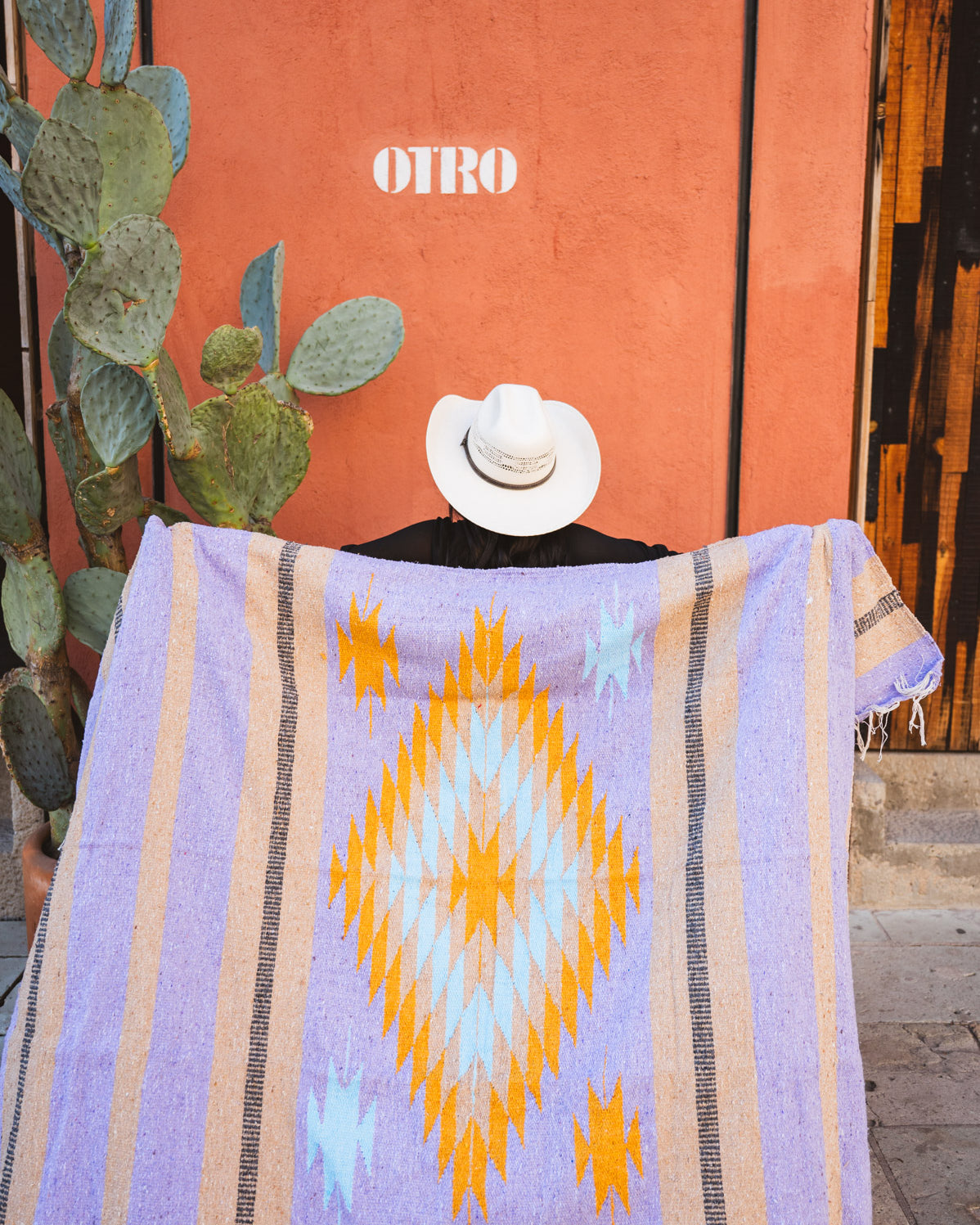 Person draped in a colorful patterned blanket with 'OTRO' branding against an orange wall.