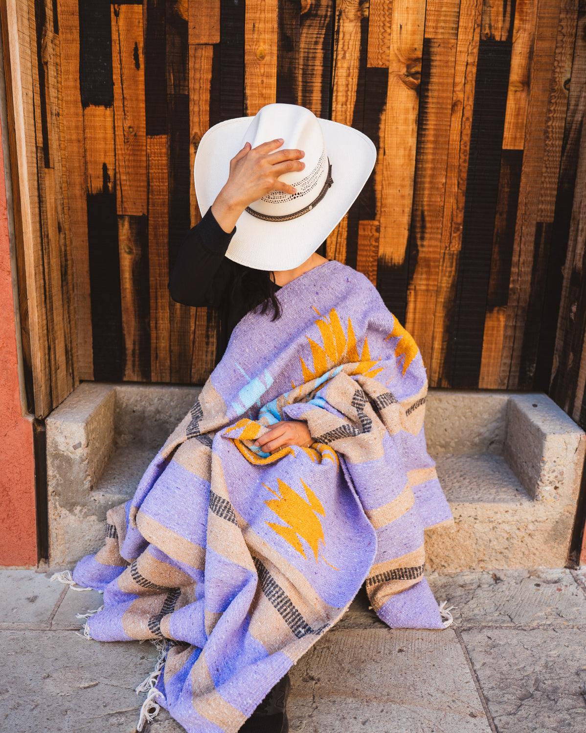 Person sitting on steps wearing a patterned blanket and white hat against a wooden wall.