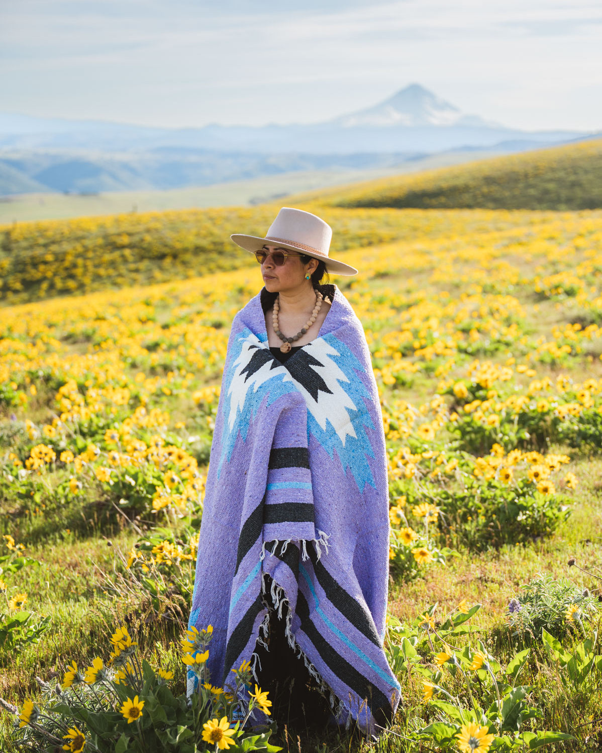 Person wrapped in a colorful blanket standing in a field of yellow flowers with mountains in the background