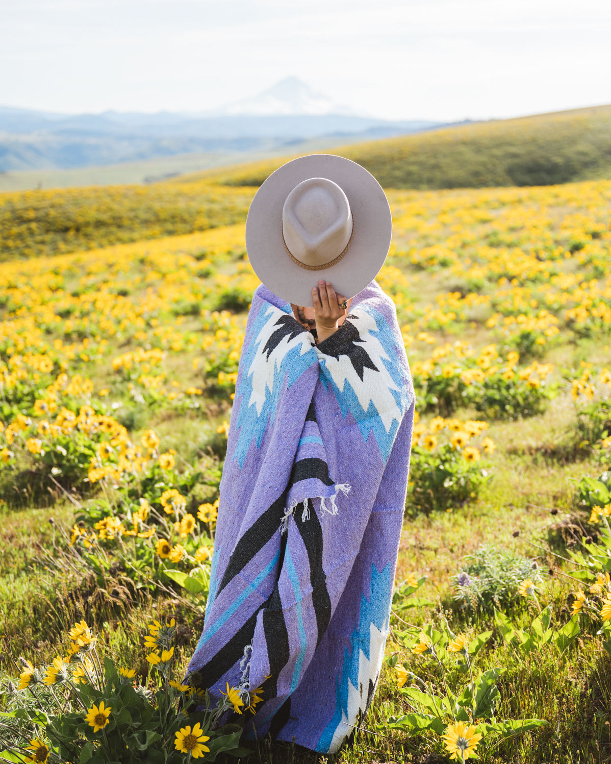Person wrapped in a colorful blanket standing in a field of yellow flowers with mountains in the background