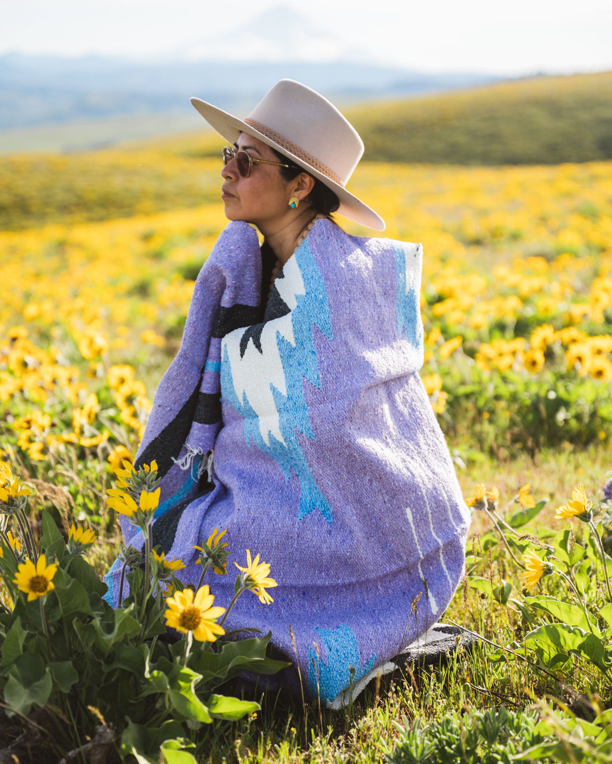 Person sitting in a field of yellow flowers wearing a colorful blanket and hat.