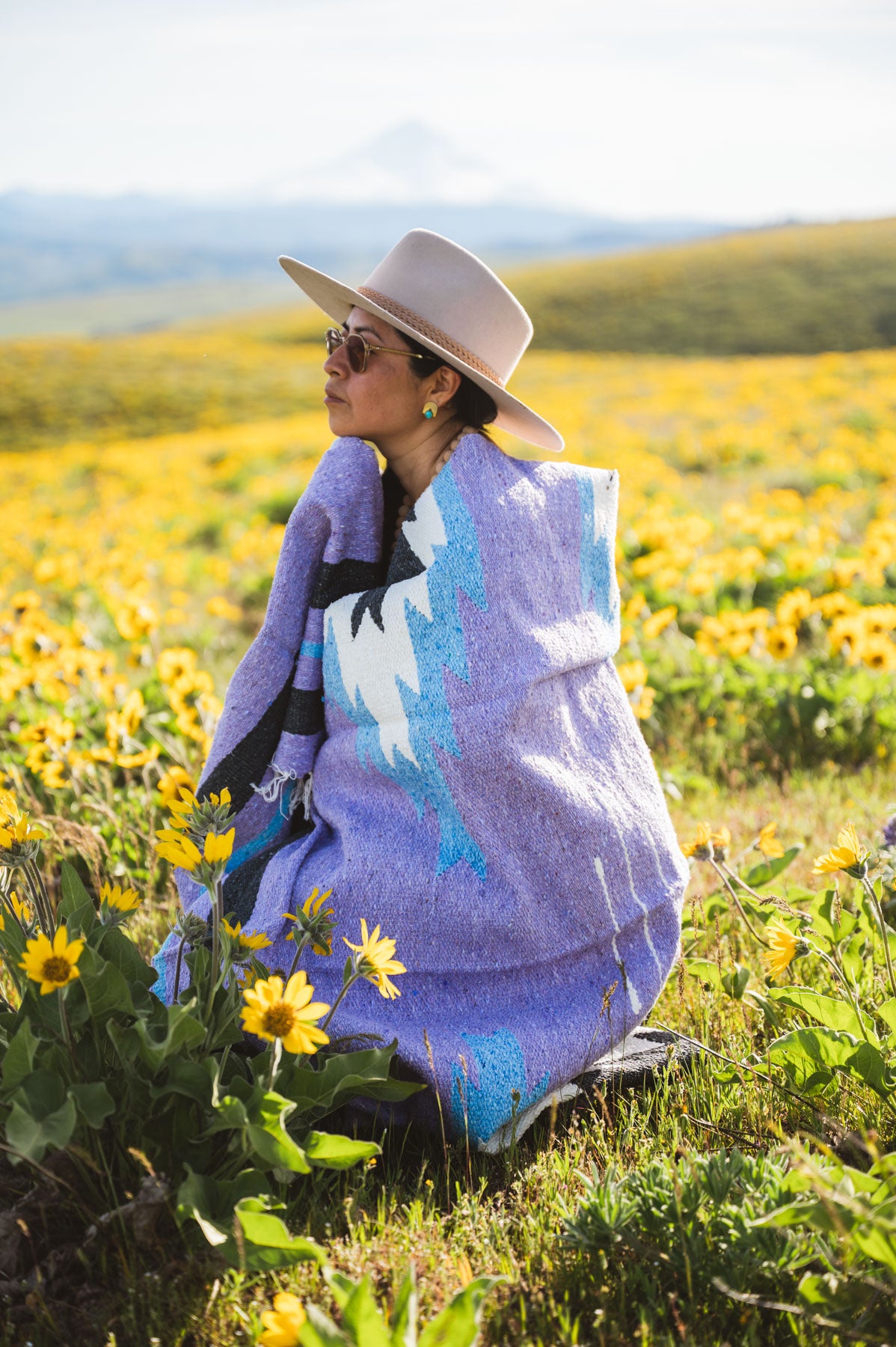 Person sitting in a field of yellow flowers wearing a colorful blanket and hat.