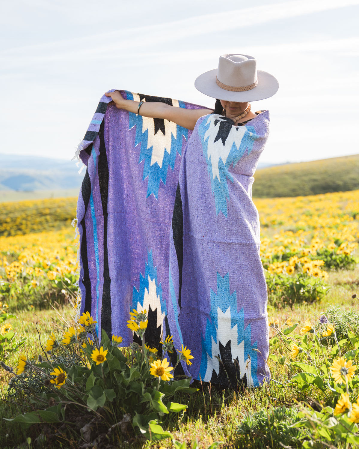 Person wrapped in a colorful patterned blanket standing in a field of yellow flowers.