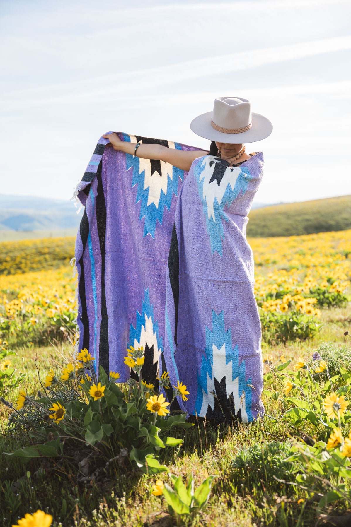 Person wrapped in a colorful patterned blanket standing in a field of yellow flowers.