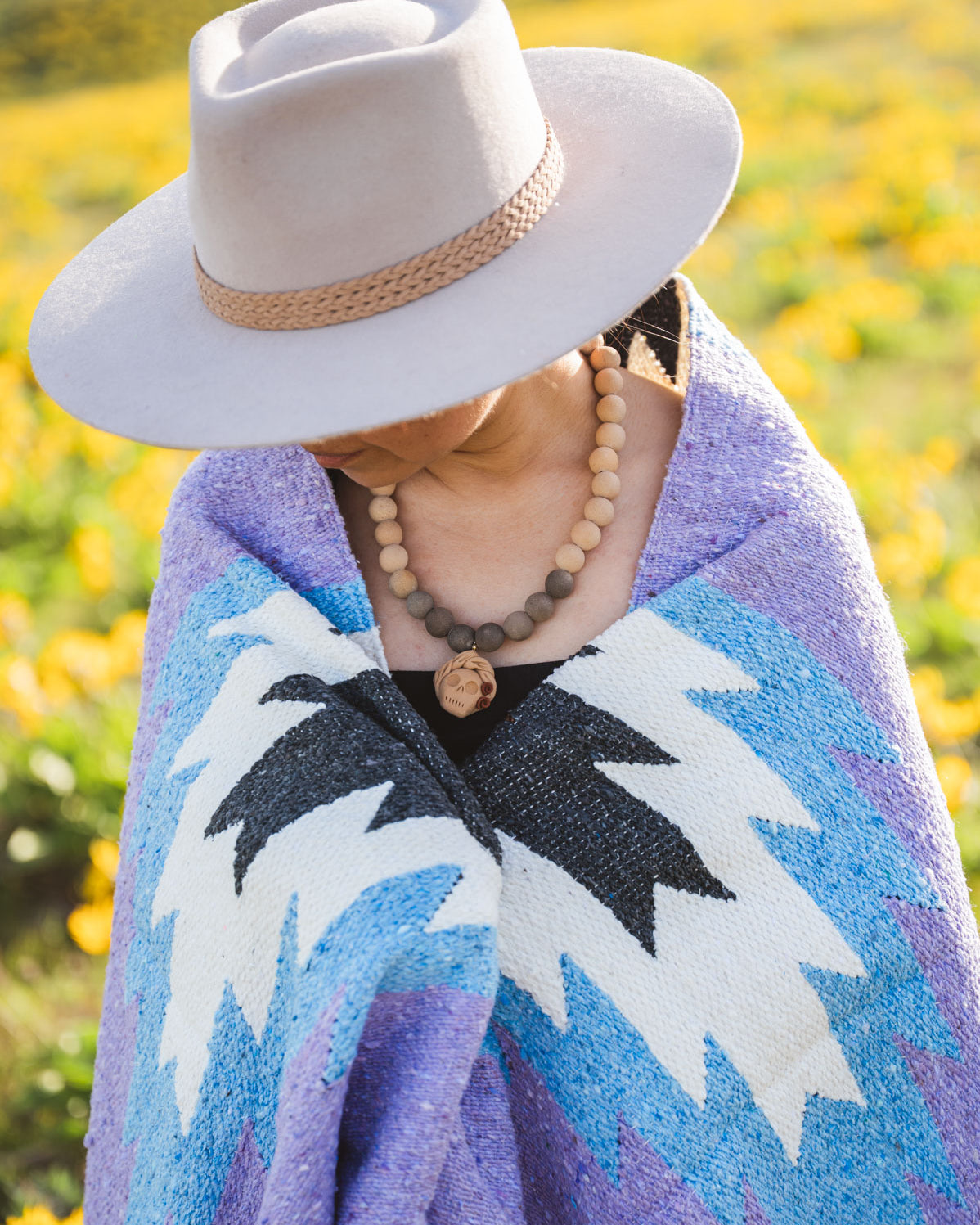 Person wearing a wide-brimmed hat and colorful patterned shawl in a field of yellow flowers.