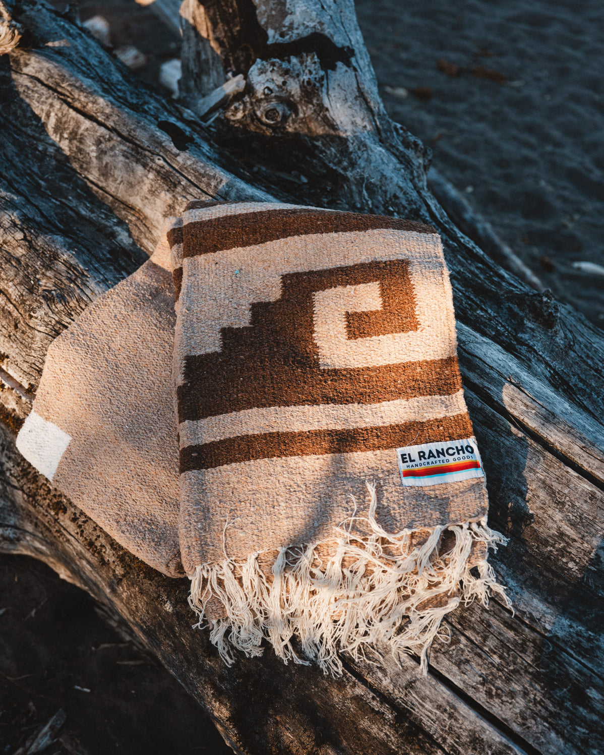 A woven throw blanket with a geometric pattern in brown and cream, draped over a piece of driftwood outdoors. The blanket has fringed edges and a small woven brand label that reads 'EL RANCHO'.