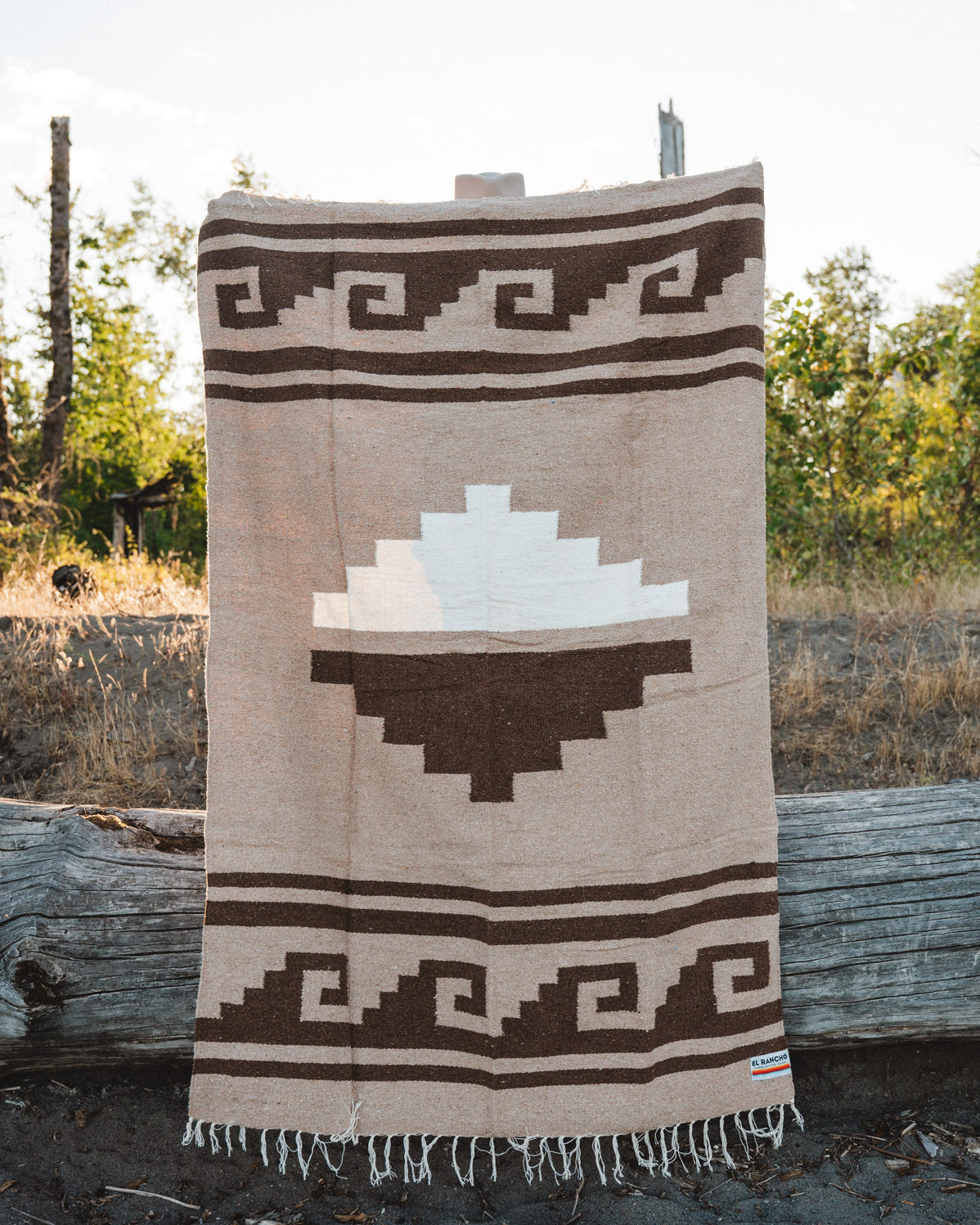 A woven blanket with a Southwestern geometric pattern in beige, brown, and white, hanging outdoors on a rustic wooden stand.