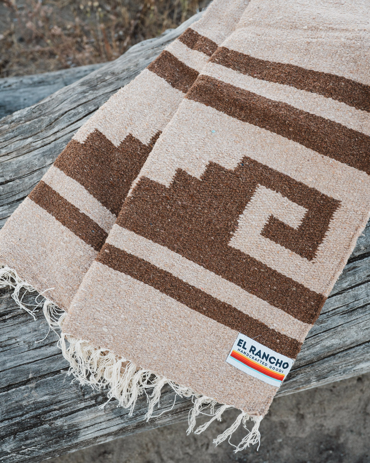 A woven blanket with a brown geometric pattern on a beige background, draped over a weathered wooden log outdoors. The blanket has fringed edges and a small woven label that reads 'EL Rancho Handwoven'.