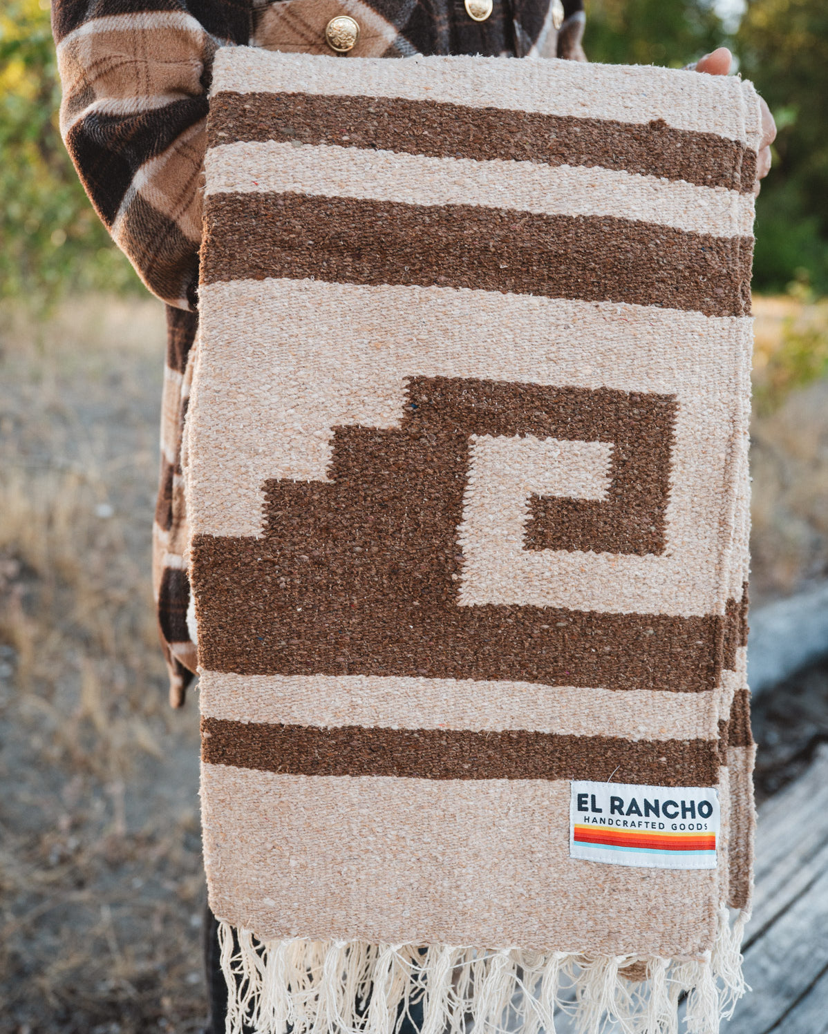 A person outdoors holds up a woven blanket with a brown and beige geometric pattern and fringe. The blanket has a visible El Rancho Handwoven label near the bottom right corner.