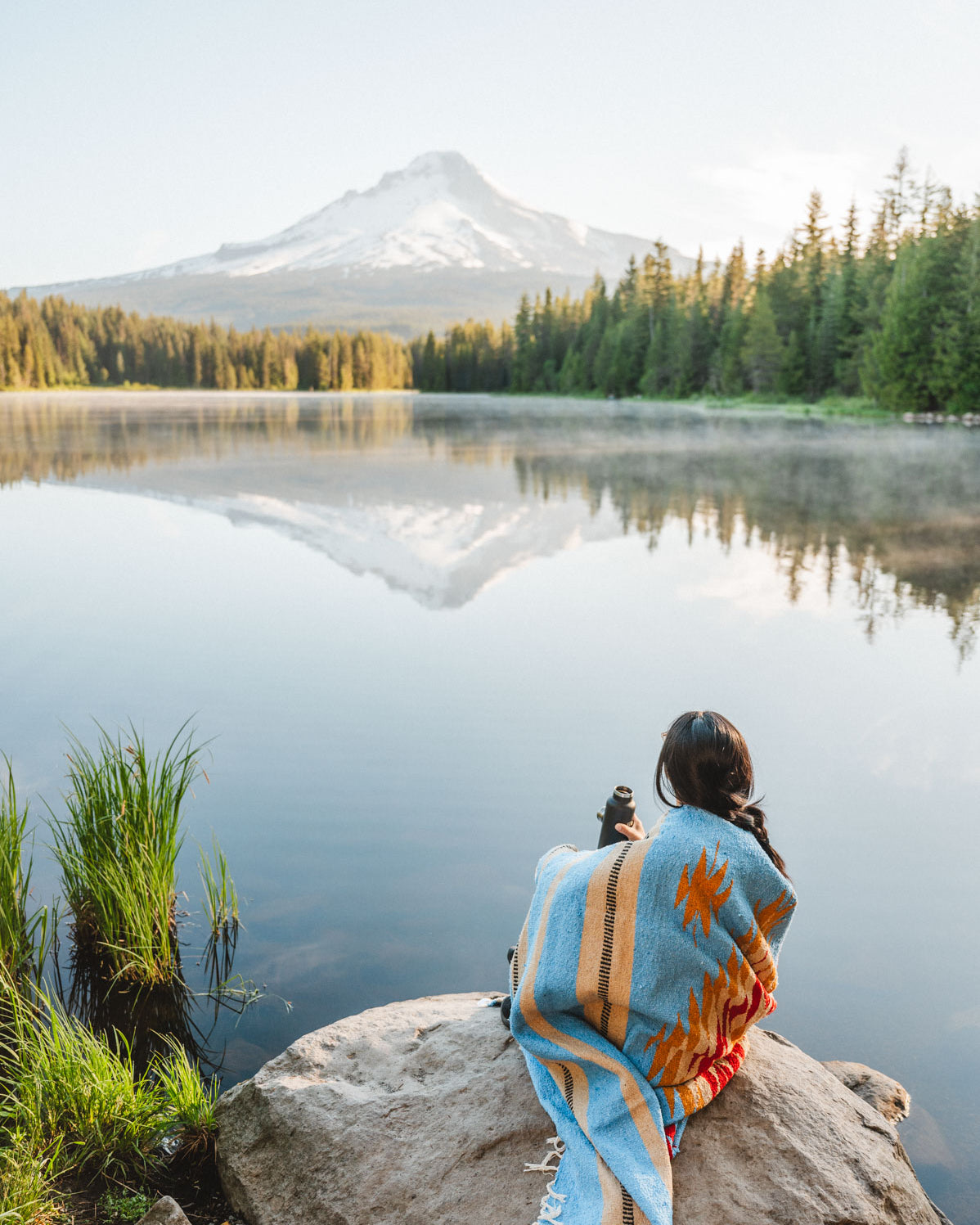 Girl wrapped in blue handwoven mexican blanket sitting on rock in front of Trillium lake looking at Mt. Hood