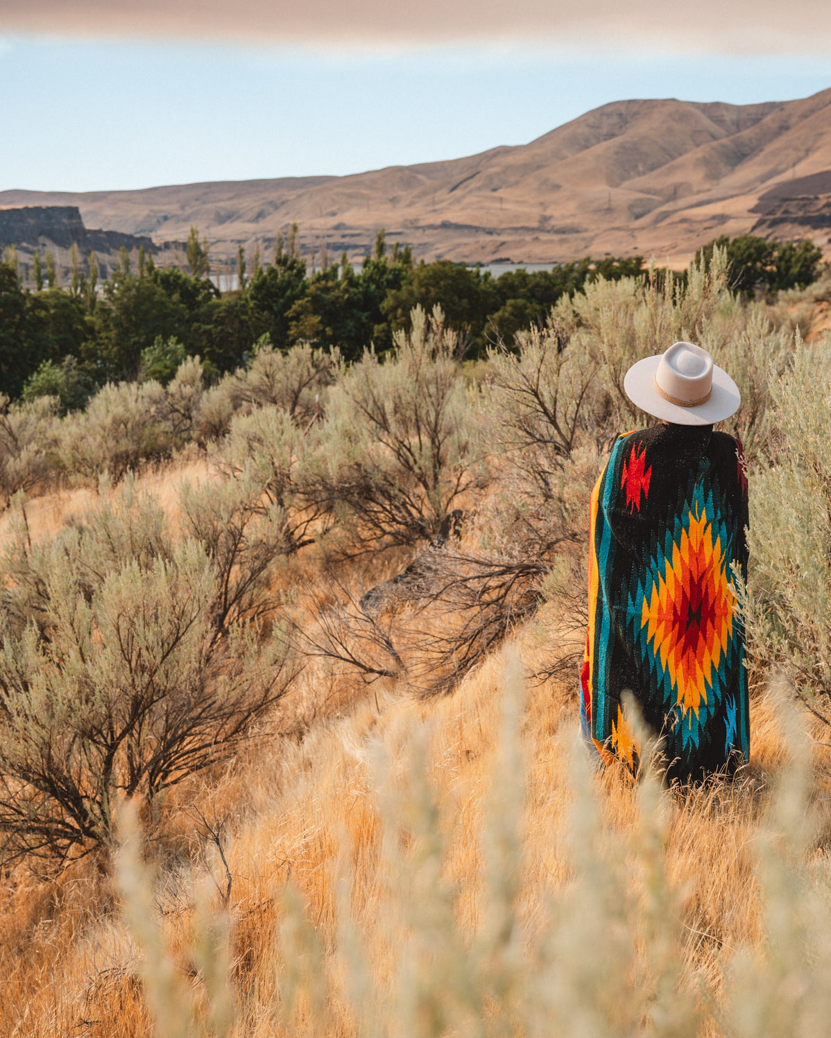 A person standing outdoors in a dry grassy landscape, viewed from behind, wearing a wide-brim white hat and a colorful geometric patterned blanket or poncho with red, orange, yellow, green, and blue motifs.