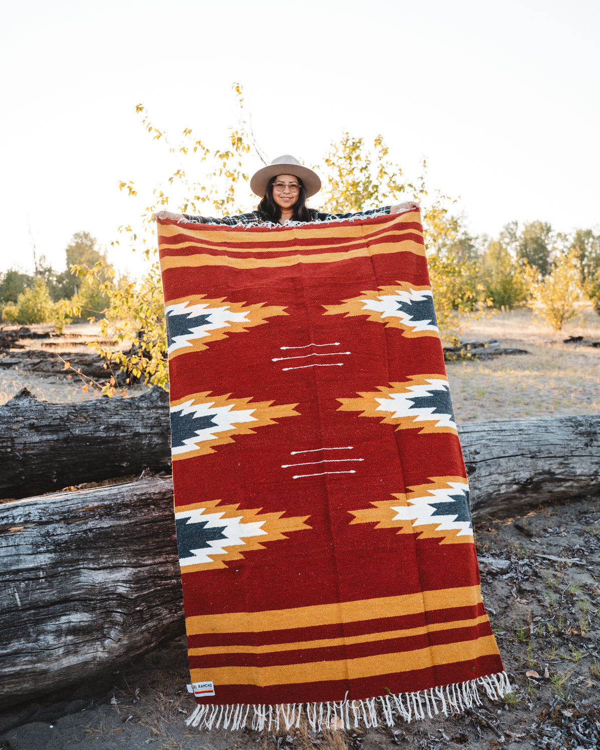 Person holding a large relampago patterned mexican blanket outdoors with trees and logs in the background