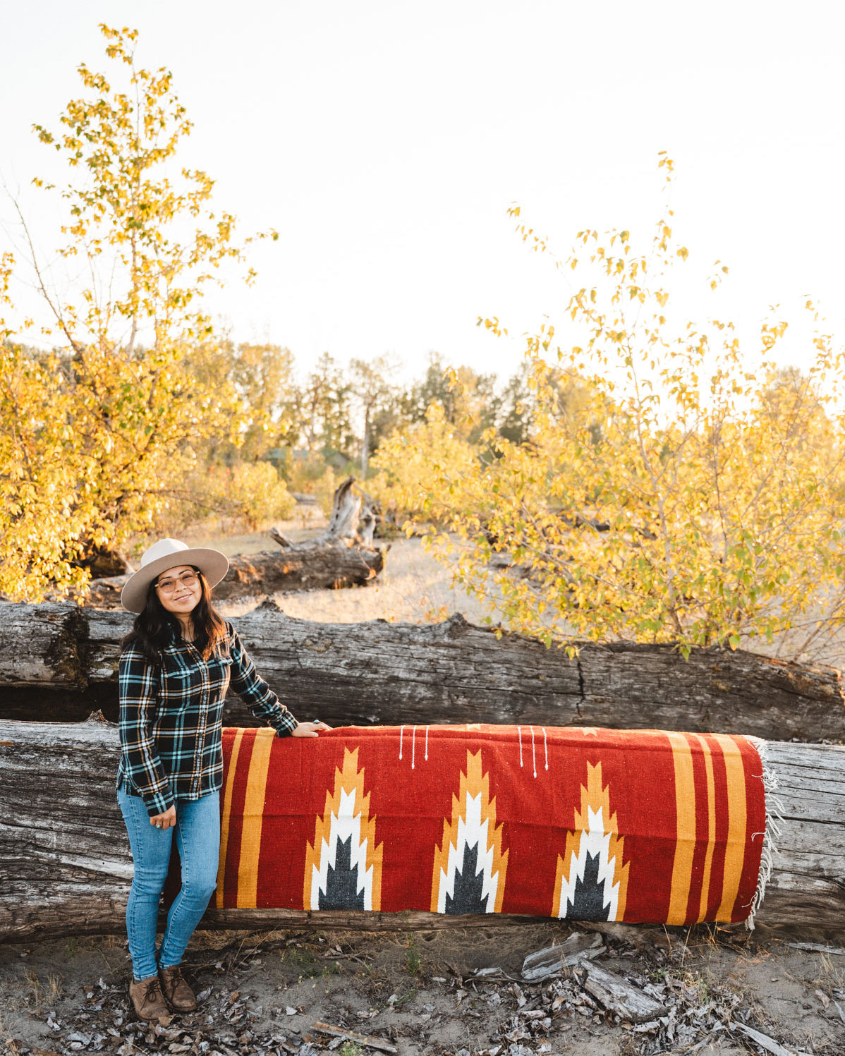Person sitting on a large relampago patterned mexican blanket outdoors with trees and logs in the background