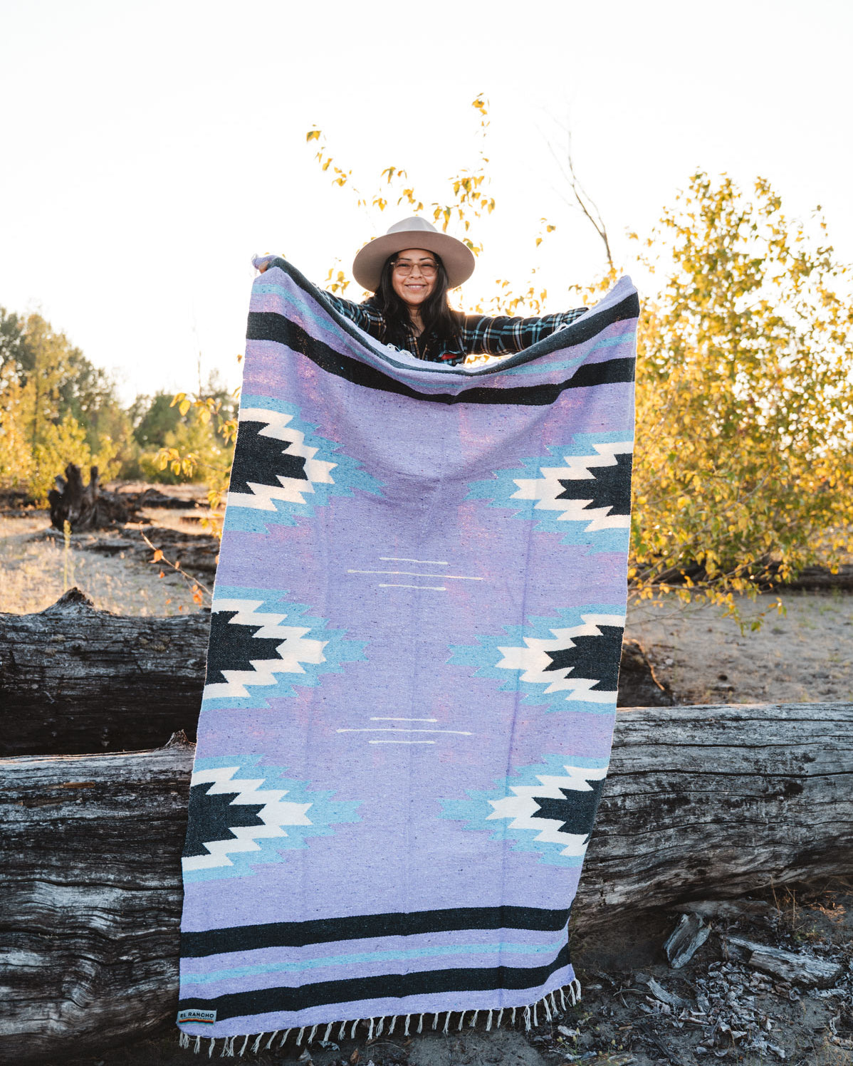 Person holding a large relampago patterned mexican blanket outdoors with trees and logs in the background