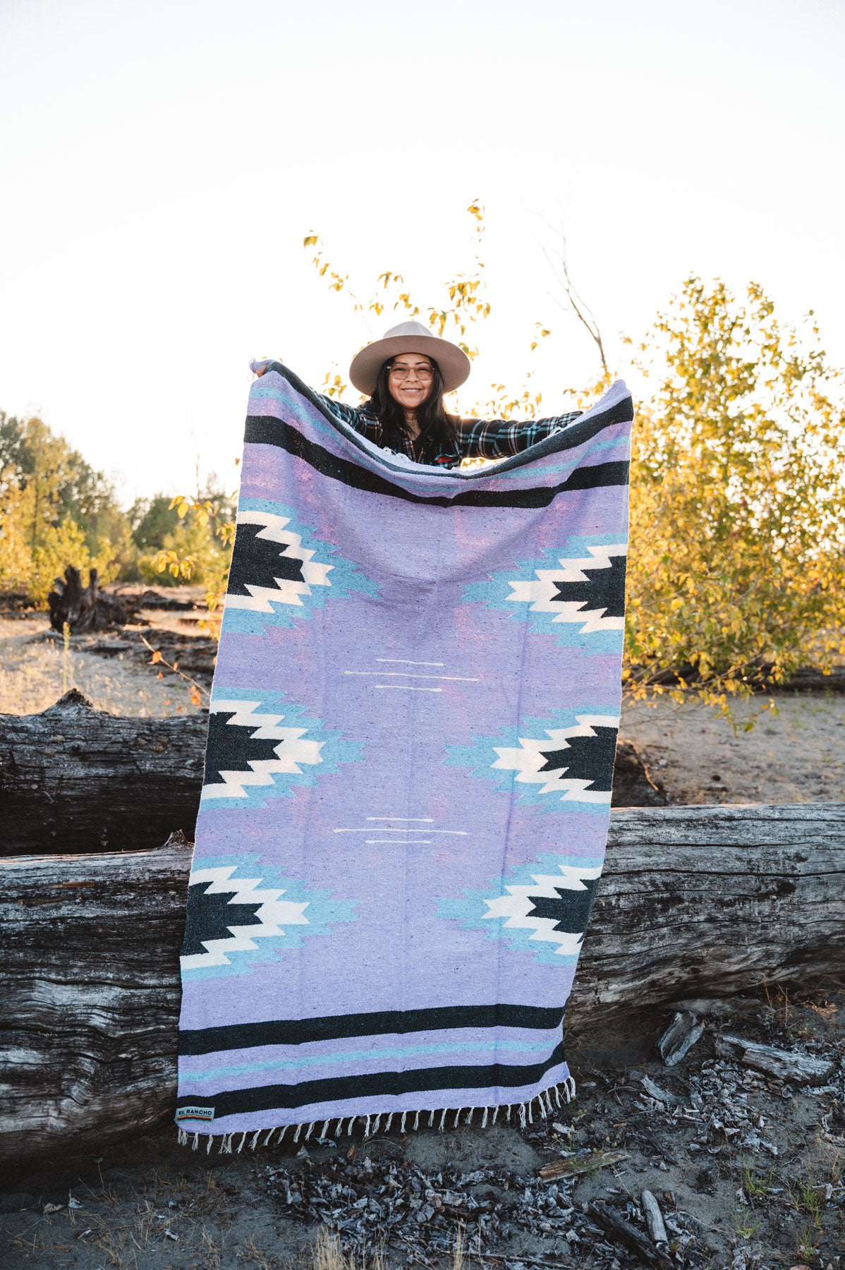 Person holding a large relampago patterned mexican blanket outdoors with trees and logs in the background