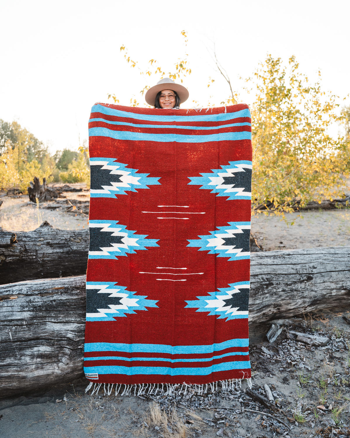 Person holding a large relampago patterned mexican blanket outdoors with trees and logs in the background