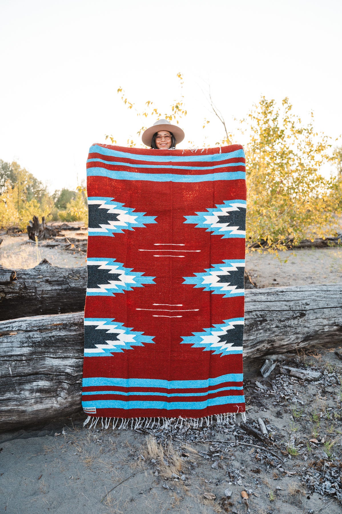 Person holding a large relampago patterned mexican blanket outdoors with trees and logs in the background
