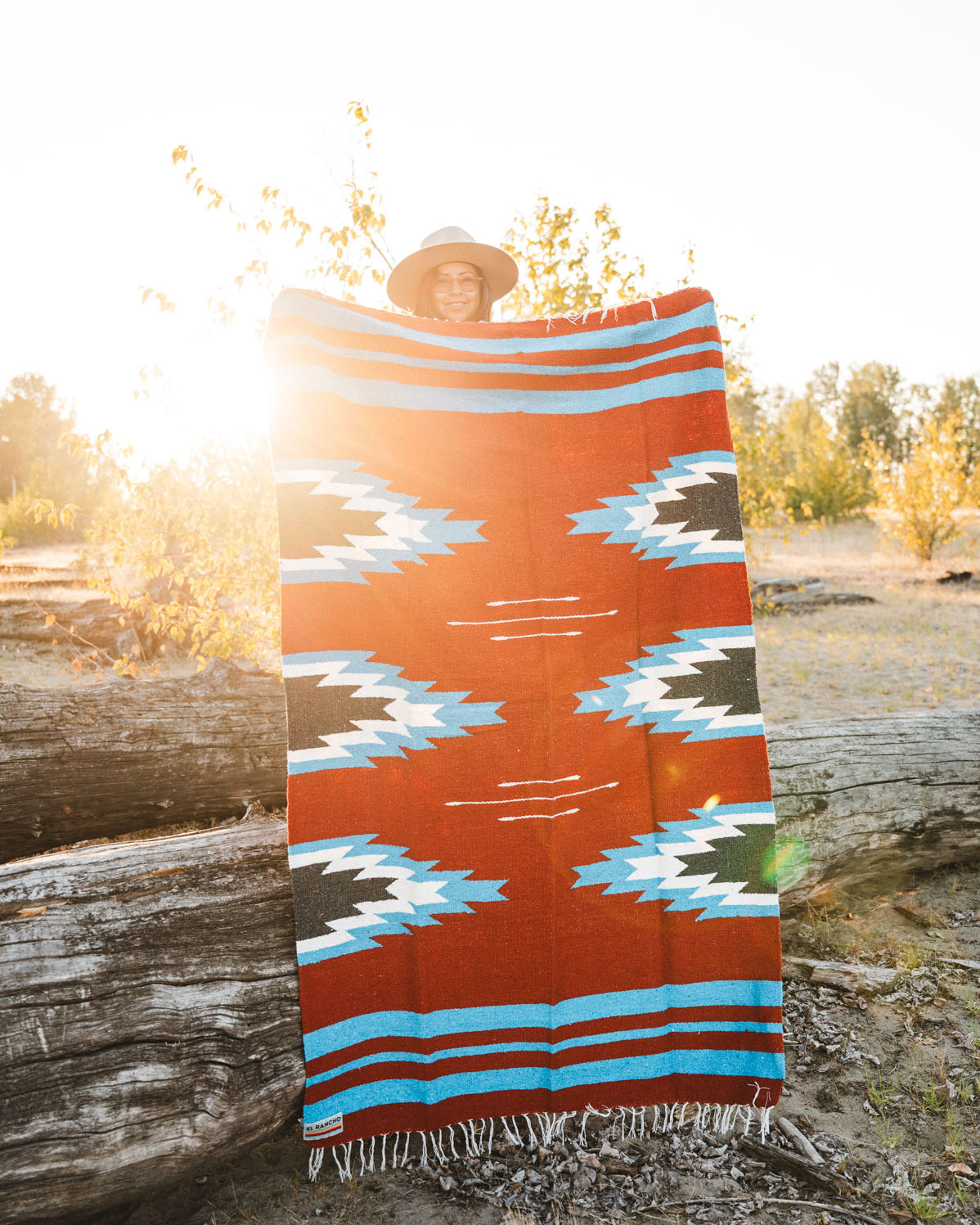 Person holding a large relampago patterned mexican blanket outdoors with trees and logs in the background