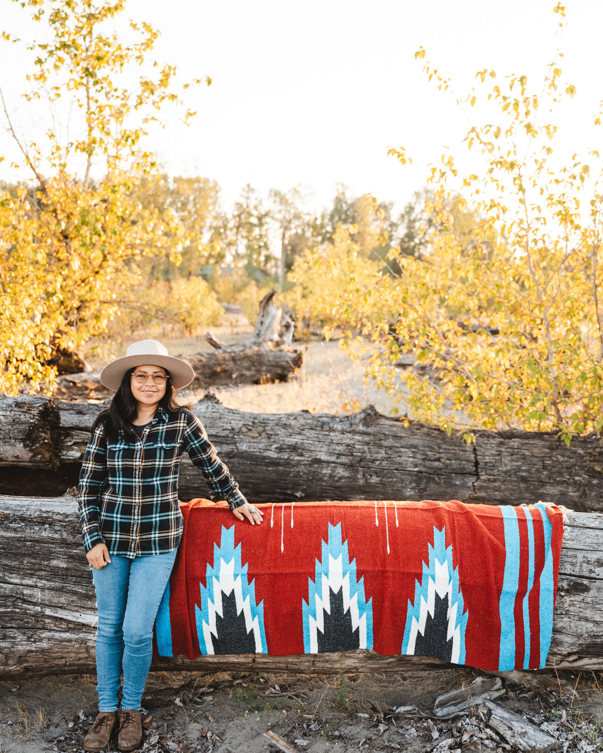 Person sitting next to a large relampago patterned mexican blanket outdoors with trees and logs in the background