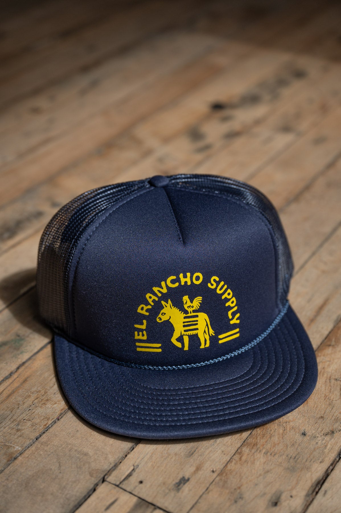A navy blue trucker hat with yellow embroidered text reading 'EL RANCHO SUPPLY' and a donkey logo, resting on a rustic wooden floor.