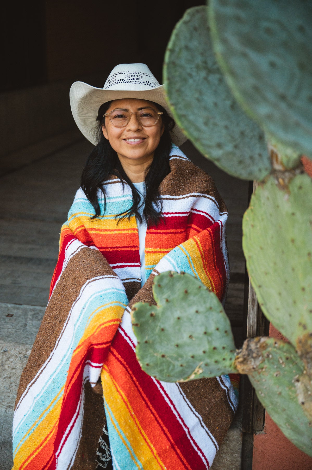 Person wearing a colorful poncho and cowboy hat, standing next to a cactus plant.