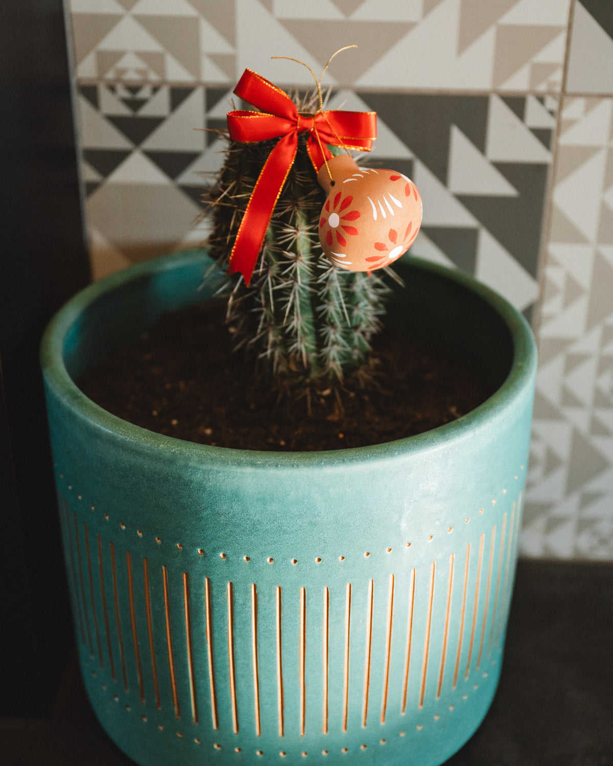 Decorative oval christmas decoration with floral pattern and red ribbon hanging on a cactus plant.