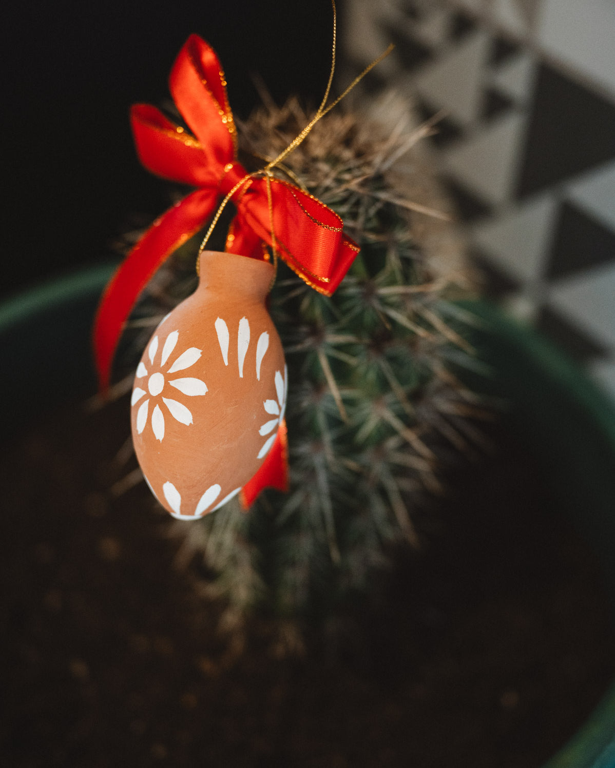 Decorative oval christmas decoration with floral pattern and red ribbon hanging on a cactus plant.