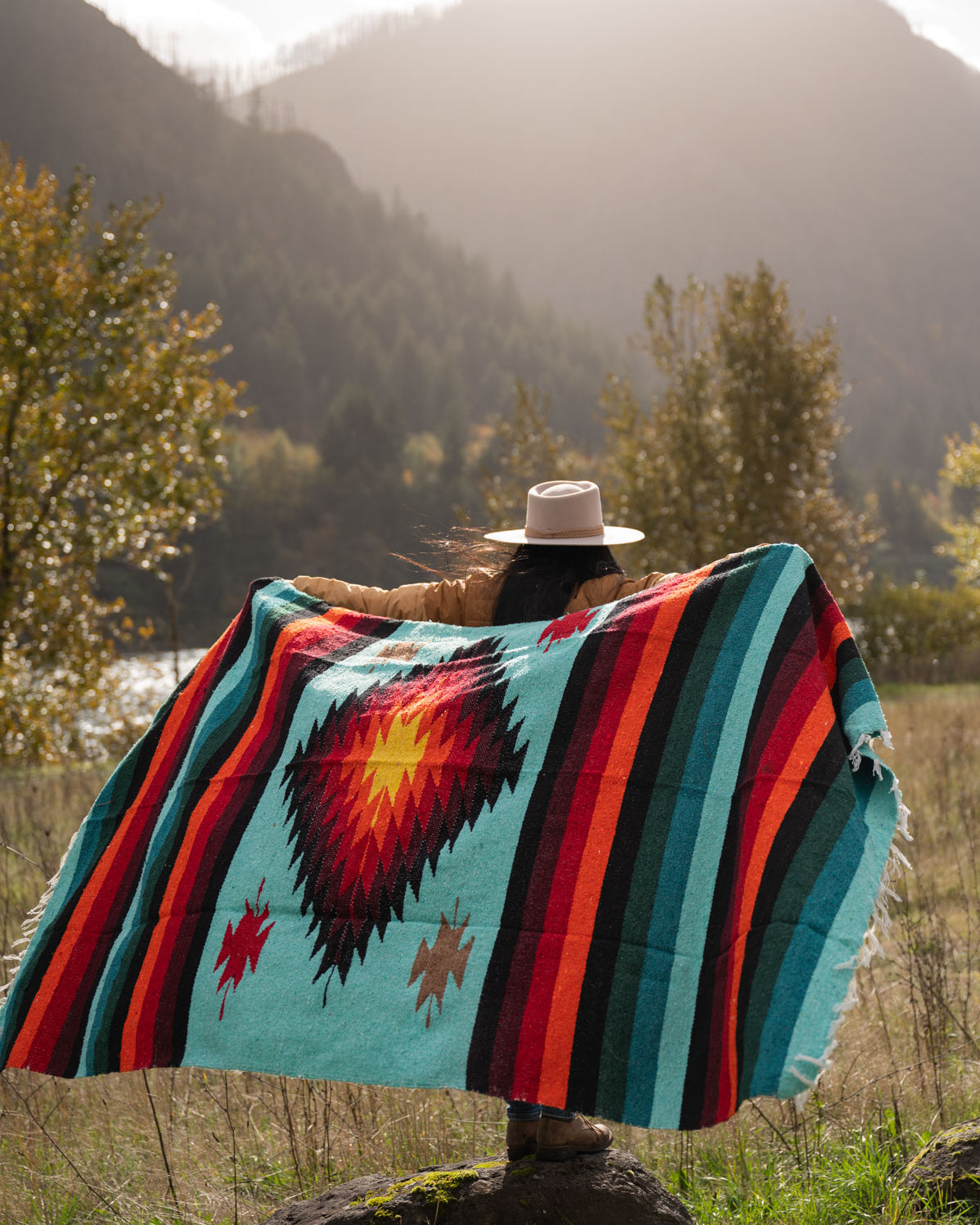 Woman wearing a cowboy hat holding a teal and red handwoven mexican diamond blanket in front of sunset in the mountains